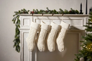 Four white knitted Christmas stockings with cable pattern hanging on a white fireplace mantle
