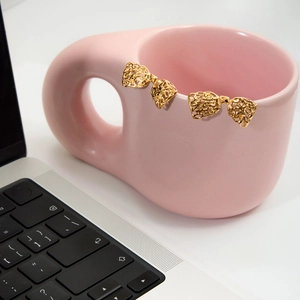 Golden titanium steel bow earrings with irregular texture hanging on a pink cup edge