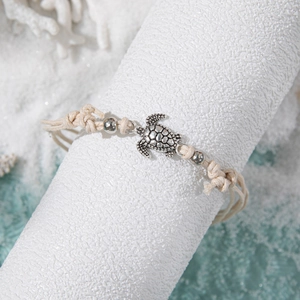 Beige woven beach anklet with silver sea turtle charm displayed on white sand