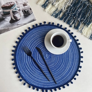 Blue round hand-woven cotton coaster with small pompoms on white desktop setting