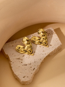 Close-up of golden heart earrings with irregular molten texture on a beige stone surface