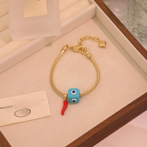 Close-up of golden snake chain bracelet featuring blue evil eye bead and red pepper charm