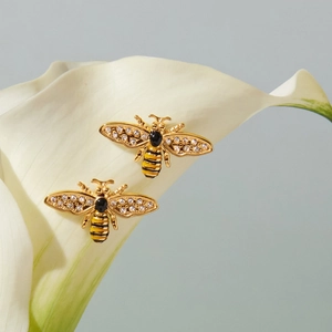 Detailaufnahme der goldenen Bienen-Ohrringe mit Kristallen auf weißen Lilienblättern