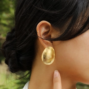Close-up of a model wearing large gold-toned textured oval stud earrings outdoors