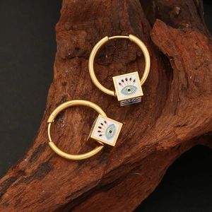 Gold hoop earrings featuring cube evil eye pendants on textured dark wood background