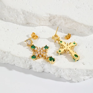 Gold-plated cross stud earrings with green zircon and pearl accents on stone texture