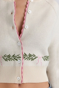 Close-up of cream knit cardigan with pink wavy trim and green leaf embroidery