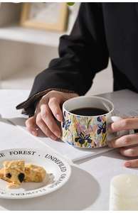 Close-up of a floral ceramic coffee cup with a large handle held by hands in a cozy setting