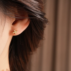 Close-up of a minimalist golden cross stud earring worn by a model with soft lighting