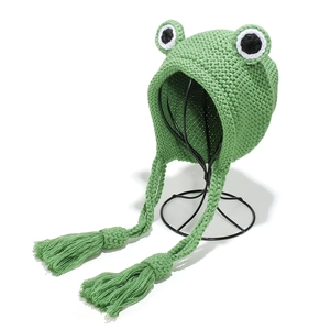 Green knit frog beanie with ear flaps and large eye detail shown on a display stand