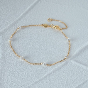 Detalle de una pulsera de cadena dorada ajustable con pequeñas perlas blancas sobre gris
