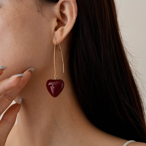 Detail view of model wearing a long gold earring with a glossy dark red heart pendant