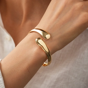 Close-up of minimalist teardrop-shaped open gold bangle on model's wrist wearing white linen