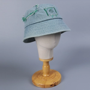 Light blue denim bucket hat with green embroidery and frayed edges on grey mannequin head