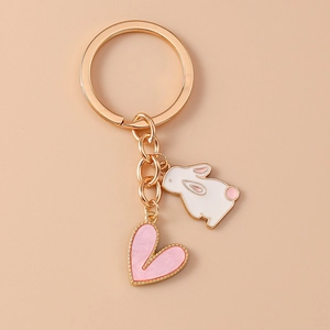 Detail showing gold keychain with white rabbit and pink heart charm on beige backdrop