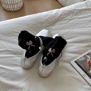 Flat lay of silver square-toe shoes with bow-embroidered socks on a soft white bed
