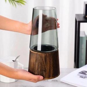 Close-up of tall smoke gray glass vase with dark wood base held by hands in a room