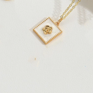 Detail of a gold necklace with a white enamel square pendant featuring a 3D floral motif