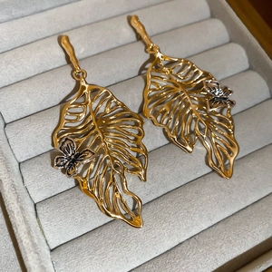 Close-up of golden hollow leaf earrings with silver butterfly accents on grey velvet pad