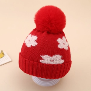 Close-up of red winter knit baby hat with white flower accents and a large plush pom-pom