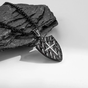 Black shield necklace with woven texture and silver cross pendant on stone surface