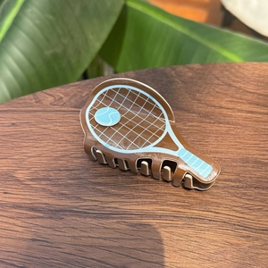 Pince à cheveux en acétate forme raquette de tennis avec détail balle bleue sur fond bois