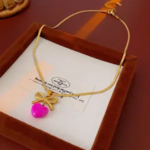Collier en acier titane avec nœud doré et pendentif cœur rose sur support bois