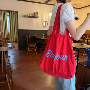Red canvas drawstring tote bag with blue Bonjour lettering shown in a cafe setting