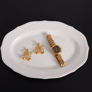 Elegant gold bow earrings and rectangular watch on a white ceramic dish