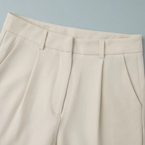 Macro detail of high-waisted cream trousers showing double pleats and belt loops