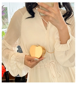 Detail of a golden heart-shaped acrylic evening bag held by a model in a white dress