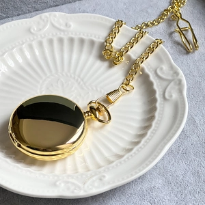 Polished gold vintage pocket watch with metal chain set on a white embossed ceramic plate