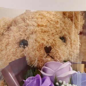 Detail of plush khaki teddy bear with purple and blue soap flower roses in a clear gift box