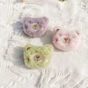 Three cute plush bear head pins in purple, pink, and green on a soft white blanket