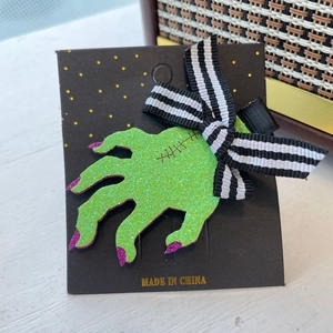 Glittery green zombie hand hair clip with purple tips and a striped bow on card