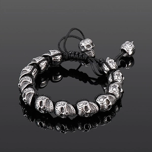 Close-up of silver skull charms on a black braided cord bracelet over a dark mirror