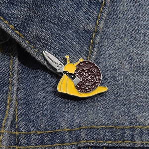 Macro detail of a cute cartoon snail enamel pin with a tiny knife on denim jacket
