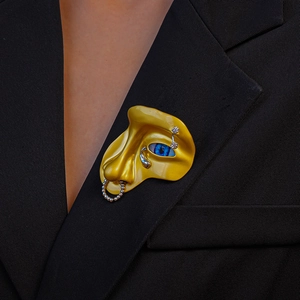 Détail macro d'une broche visage dorée surréaliste avec oeil bleu et cristaux sur veste