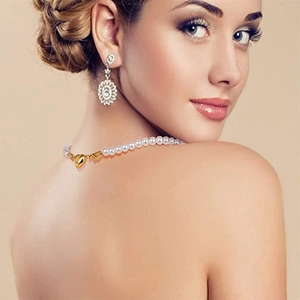 Close-up of pearl necklace with gold heart clasp and diamond earrings worn by model