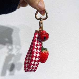 Close-up of a strawberry keychain with red bell and houndstooth woven strap held by hand