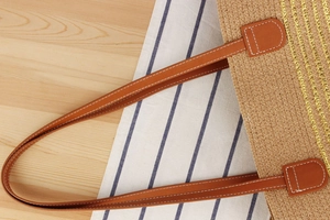 Close-up of woven straw tote bag with brown leather handles and gold metallic stripes