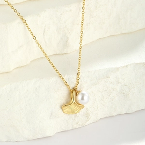 Detail of a gold ginkgo leaf necklace with a white pearl on a textured stone base