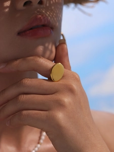 Golden oval statement ring on finger with summer straw hat shadows under blue sky