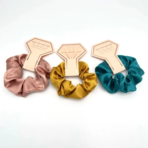 Three satin scrunchies in rose gold, gold and blue on diamond-shaped card packaging