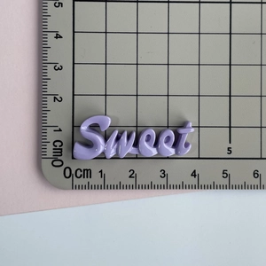 Détail du charm Sweet en résine violette montrant des dimensions de 3,5 cm sur un tapis
