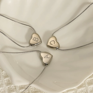 Three stainless steel heart letter necklaces on a shell dish with snake chain