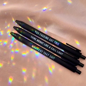 Detailed view of matte black funny pens with holographic text on pink silk fabric