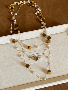 Detail of layered gold necklace featuring tiger eye beads and pearls on a beige tray