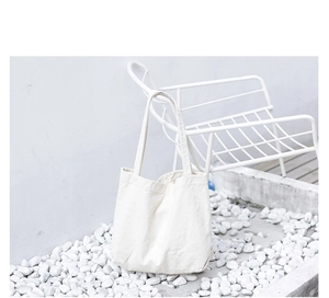 Minimalist white canvas tote bag resting on pebbles with a white metal chair backdrop