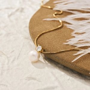 Detailed close-up of a gold necklace pendant featuring a pearl and zircon on a textured mat